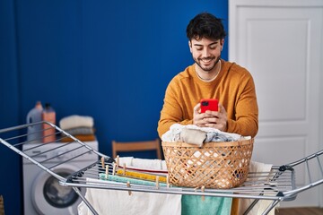 Sticker - Young hispanic man using smartphone hanging clothes on clothesline at laundry room