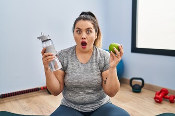 Wall Mural - Young hispanic woman holding green apple and bottle of water afraid and shocked with surprise and amazed expression, fear and excited face.