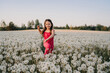 Charming girl in pink jumpsuit standing in field surrounded by white dandelions and smiling while looking at blossoms.
