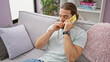 Young hispanic man speaking on the phone sitting on sofa yawning at home