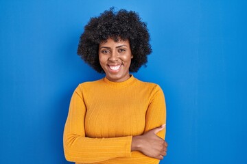 Poster - Black woman with curly hair standing over blue background happy face smiling with crossed arms looking at the camera. positive person.