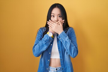 Sticker - Young asian woman standing over yellow background shocked covering mouth with hands for mistake. secret concept.