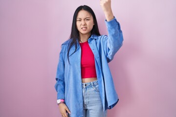 Sticker - Young asian woman standing over pink background angry and mad raising fist frustrated and furious while shouting with anger. rage and aggressive concept.