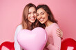 Charming long-haired girls smile and hug each other holding a heart on a pink background