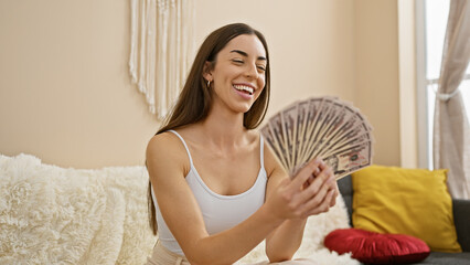 Canvas Print - Attractive young hispanic woman smiling, flaunting her wealth as she holds dollar banknotes, sitting in the comfort of her home's lavish living room sofa.