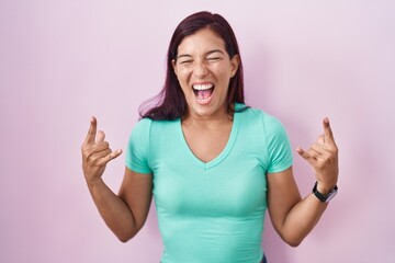 Poster - Young hispanic woman standing over pink background shouting with crazy expression doing rock symbol with hands up. music star. heavy music concept.