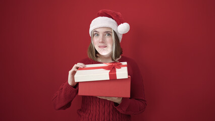 Young blonde woman wearing christmas hat unpacking gift over isolated red background