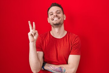 Sticker - Young hispanic man standing over red background smiling with happy face winking at the camera doing victory sign with fingers. number two.