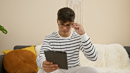 Canvas Print - Handsome young hispanic man comfortably engrossed, using touchpad on the sofa in his relaxing home indoors, reflecting a relaxed lifestyle amidst technology