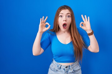 Wall Mural - Redhead woman standing over blue background looking surprised and shocked doing ok approval symbol with fingers. crazy expression