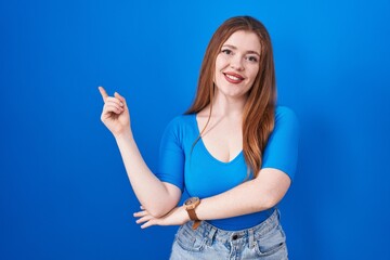 Wall Mural - Redhead woman standing over blue background with a big smile on face, pointing with hand finger to the side looking at the camera.