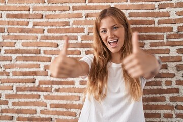 Sticker - Young caucasian woman standing over bricks wall approving doing positive gesture with hand, thumbs up smiling and happy for success. winner gesture.