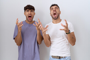 Wall Mural - Homosexual gay couple standing over white background crazy and mad shouting and yelling with aggressive expression and arms raised. frustration concept.