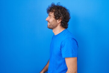 Poster - Hispanic young man standing over blue background looking to side, relax profile pose with natural face and confident smile.