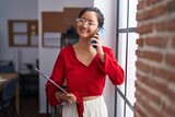 Fototapeta  - Young chinese woman business worker talking on smartphone holding clipboard at office