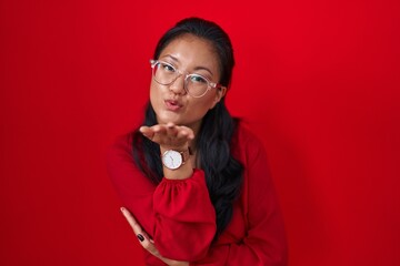 Poster - Asian young woman standing over red background looking at the camera blowing a kiss with hand on air being lovely and sexy. love expression.