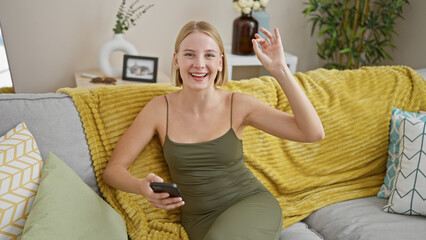 Wall Mural - Young blonde woman using smartphone doing ok gesture at home