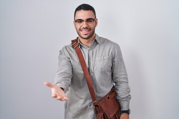 Sticker - Young hispanic man wearing suitcase smiling cheerful offering palm hand giving assistance and acceptance.