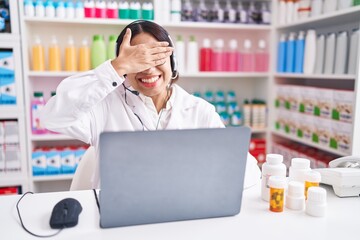 Wall Mural - Young arab woman working at pharmacy drugstore using laptop smiling and laughing with hand on face covering eyes for surprise. blind concept.