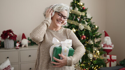 Wall Mural - Middle age woman with grey hair listening to music standing by christmas tree at home