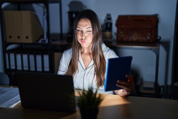 Poster - Young brunette woman working at the office at night puffing cheeks with funny face. mouth inflated with air, crazy expression.