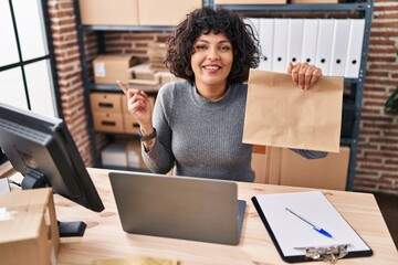 Poster - Hispanic doctor woman with curly hair working at small business ecommerce holding paper bag smiling happy pointing with hand and finger to the side