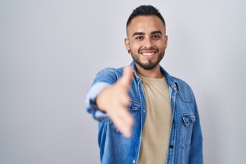 Poster - Young hispanic man standing over isolated background smiling friendly offering handshake as greeting and welcoming. successful business.