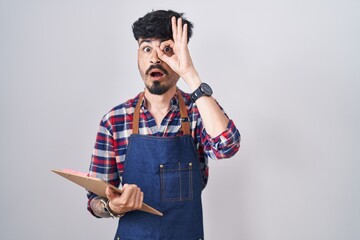 Sticker - Young hispanic man with beard wearing waiter apron holding clipboard doing ok gesture shocked with surprised face, eye looking through fingers. unbelieving expression.