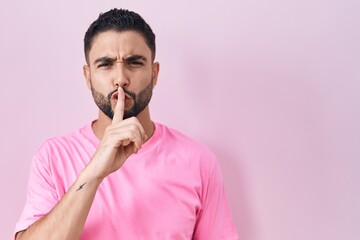 Poster - Hispanic young man standing over pink background asking to be quiet with finger on lips. silence and secret concept.