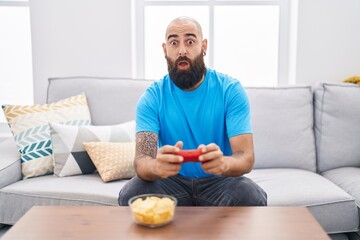 Canvas Print - Young hispanic man with beard and tattoos playing video game sitting on the sofa afraid and shocked with surprise and amazed expression, fear and excited face.