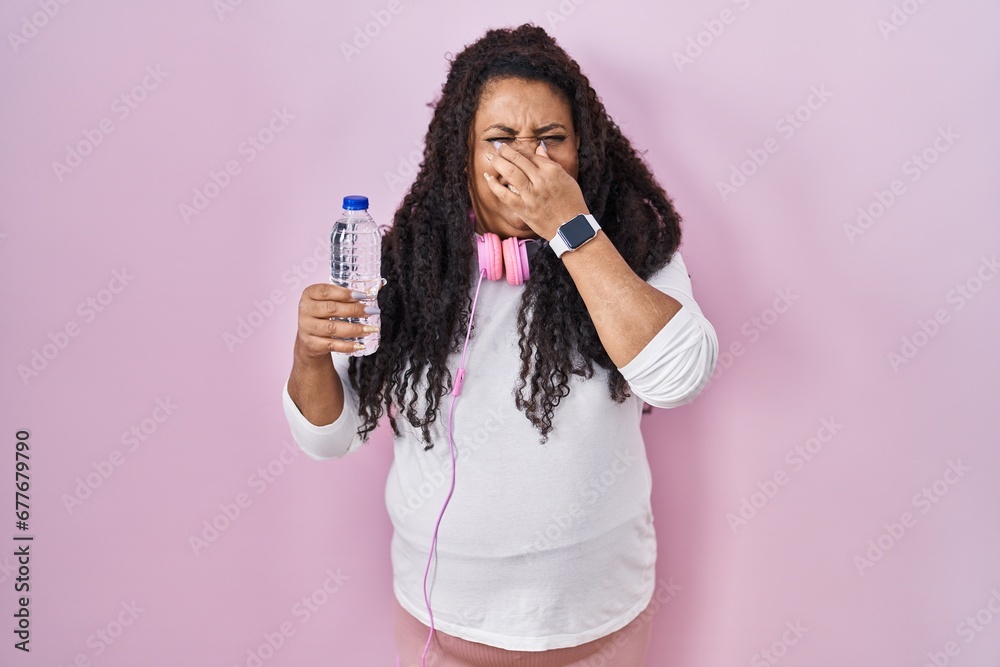 Plus size hispanic woman wearing sportswear and headphones smelling something stinky and disgusting, intolerable smell, holding breath with fingers on nose. bad smell