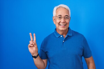 Canvas Print - Middle age man with grey hair standing over blue background smiling with happy face winking at the camera doing victory sign. number two.