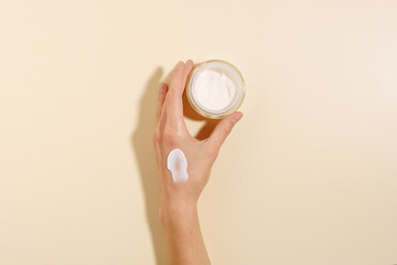 Female hand with cream smear holding glass jar of moisturizer on beige isolated background. The concept of moisturizing and nourishing the skin of the face and body, beauty products