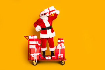 Wall Mural - Full length photo of charismatic aged santa stand market pushcart hold new year giftbox order isolated on yellow color background