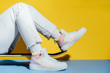 Close Up Female Legs In White Jeans And Retro Style High-top Multicolor Sport Sneakers Shoes Sitting On The Skateboard On Blue And Yellow Background. Vintage Retro Fashion Style Of 80s - 90s Vibes.