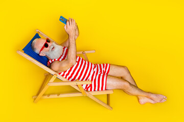 Full body photo of funny old man with white beard talking to family phone video call when lying sunbed isolated on yellow color background