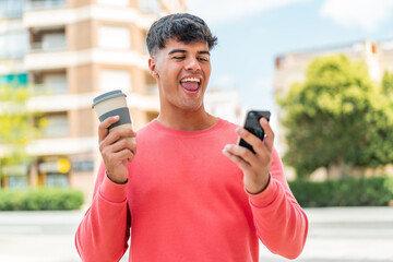 Wall Mural - Young hispanic man at outdoors using mobile phone and holding a coffee with happy expression