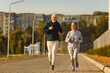 © Studio Romantic - Fit senior couple enjoying jogging workout on sunny summer morning. Old male and female athletes running on pavement, with green trees and apartment buildings in background. Sport, exercise concept