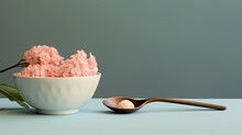  A White Bowl Filled With Pink Flowers Next To A Wooden Spoon And A Green Leaf On A Light Blue Surface.