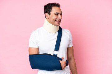 Wall Mural - Young caucasian man with broken arm and wearing a sling isolated on pink background looking side