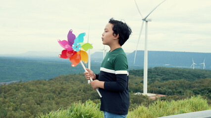 Sticker - Progressive young asian boy playing with wind turbine toy in the wind turbine farm, green field over the hill. Green energy from renewable electric wind generator.