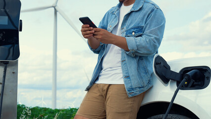 Sticker - Progressive man with his electric car, EV car recharging energy from charging station on green field with wind turbine as concept of future sustainable energy. Electric vehicle with energy generator.