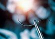 Hemostatic forceps holding half circle cutting needle and suture in front of blurry operators under the lights in operating room.