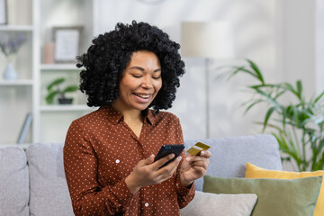 Joyful happy woman buyer online internet store, satisfied smiling sitting on the sofa in living room on house, holding a bank credit card in hands, using application on her phone, placing an order