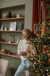 © RooM The Agency - Smiling woman standing by an illuminated Christmas tree in a stylish living room with a hot drink