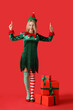 canvas print picture - Beautiful young woman in elf costume with Christmas gift boxes pointing at something on red background