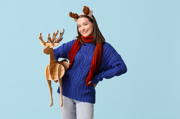 Wall Mural - Young woman in warm sweater and wooden reindeer on blue background