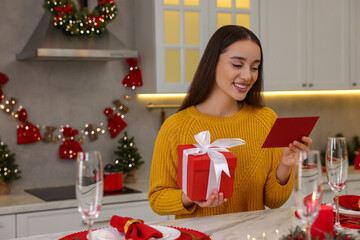 Poster - Happy young woman with Christmas gift reading greeting card at table in kitchen