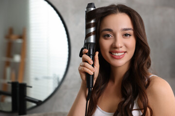 Wall Mural - Smiling woman using curling hair iron in bathroom. Space for text