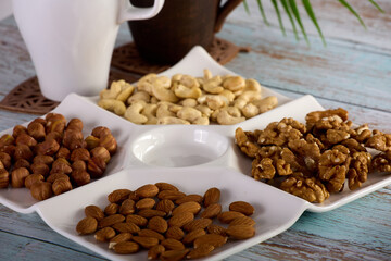 Wall Mural - Assorted dried nuts of various types are laid out in close-up on a white square plate. A mix of hazelnuts, almonds, cashews and walnuts is laid out in a white bowl standing on a wooden table